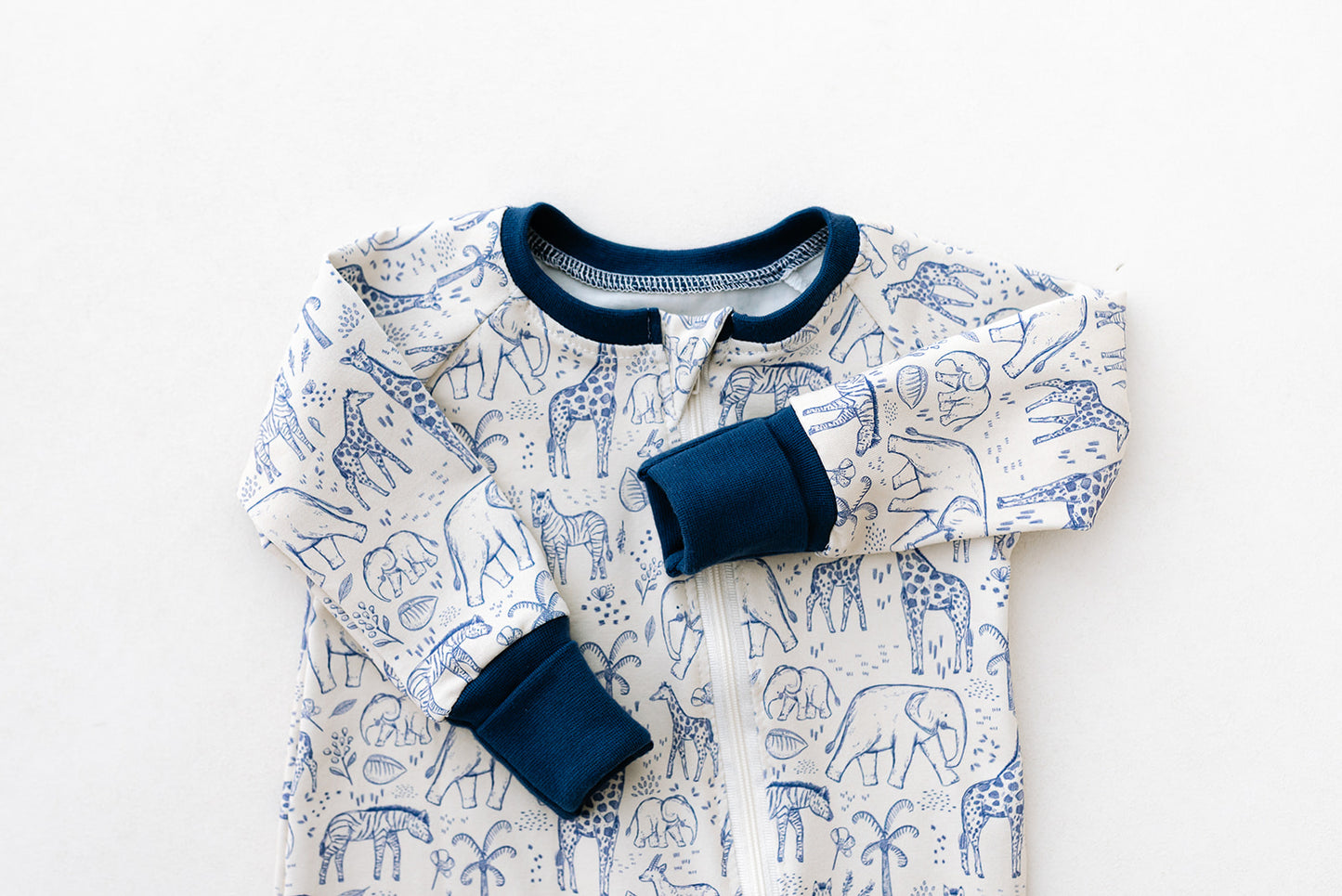 Image shows a handmade double zip baby sleepsuit in a neutral safari animal print in navy blue line artwork on an off white background with colour matching navy foldover hand and foot cuffs