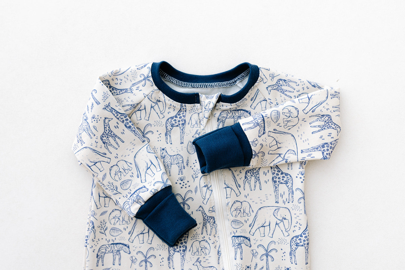 Image shows a handmade double zip baby sleepsuit in a neutral safari animal print in navy blue line artwork on an off white background with colour matching navy foldover hand and foot cuffs